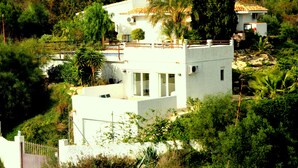 Un edificio blanco con techo plano, rodeado de abundante vegetación y una pequeña piscina.