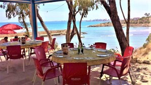 Un área de comedor al aire libre con mesas redondas y sillas rojas, con vista a una masa de agua y un horizonte urbano distante.
