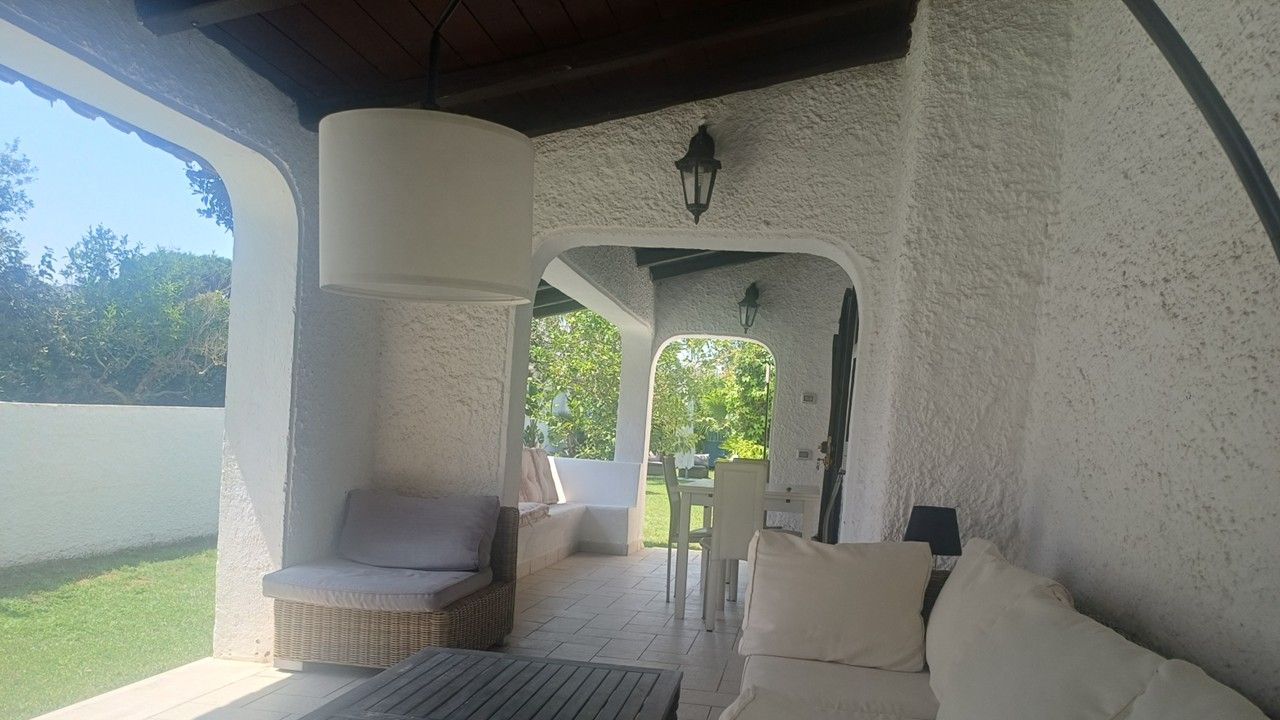 A covered patio area with a wicker chair, a white sofa, and a table. There is a view of greenery outside.