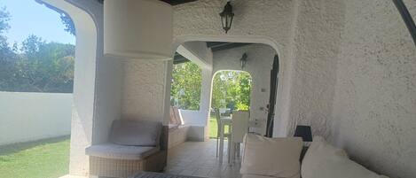 A covered patio area with a wicker chair, a white sofa, and a table. There is a view of greenery outside.