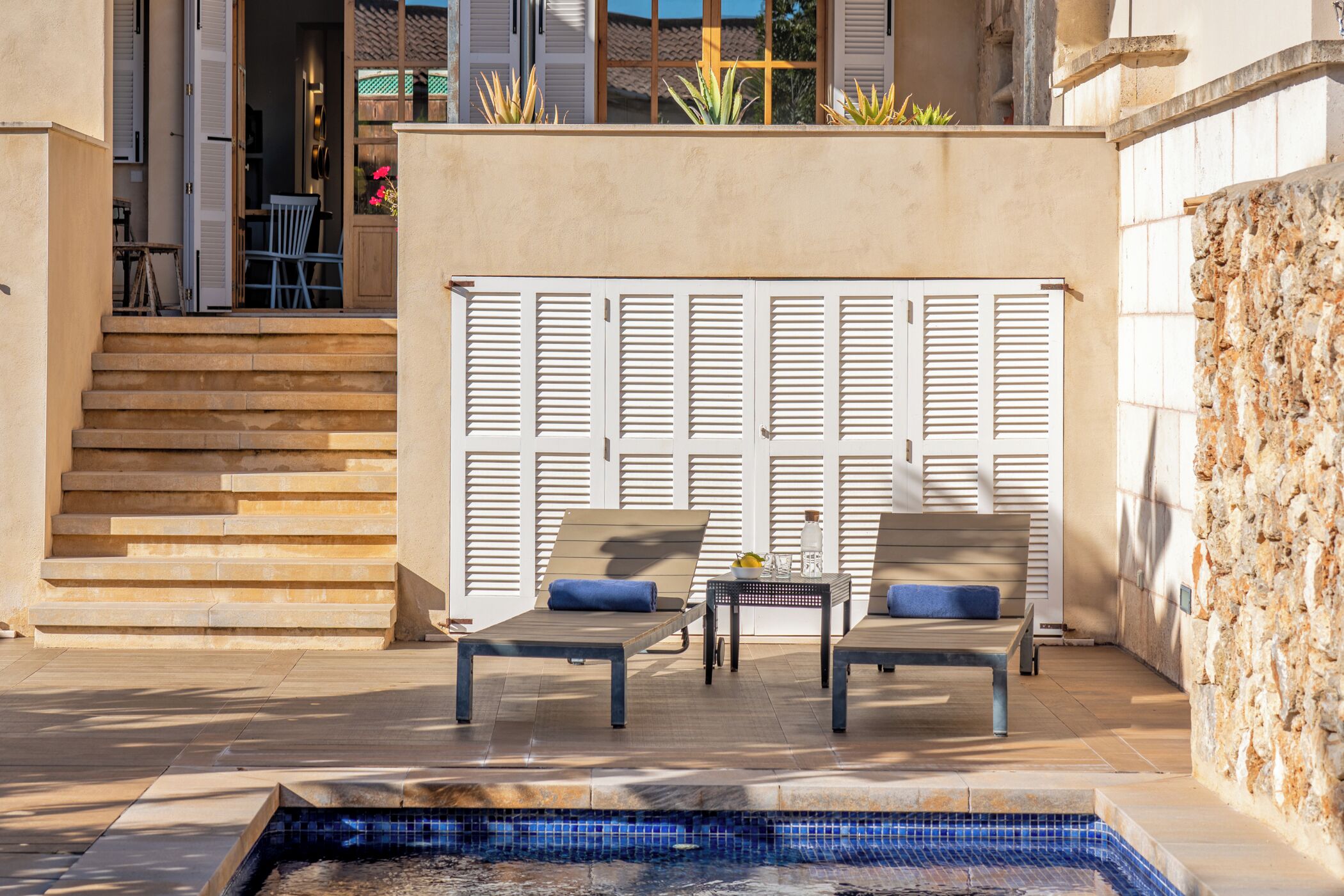A pool area with two lounge chairs and a small table, a stone wall, and a set of stairs leading to a door.