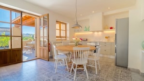A modern kitchen with white cabinetry, a dining area with a wooden table and white chairs, and a large window with a view of greenery. - Casa Son Bordoi, private pool & tranquility (Llubí)