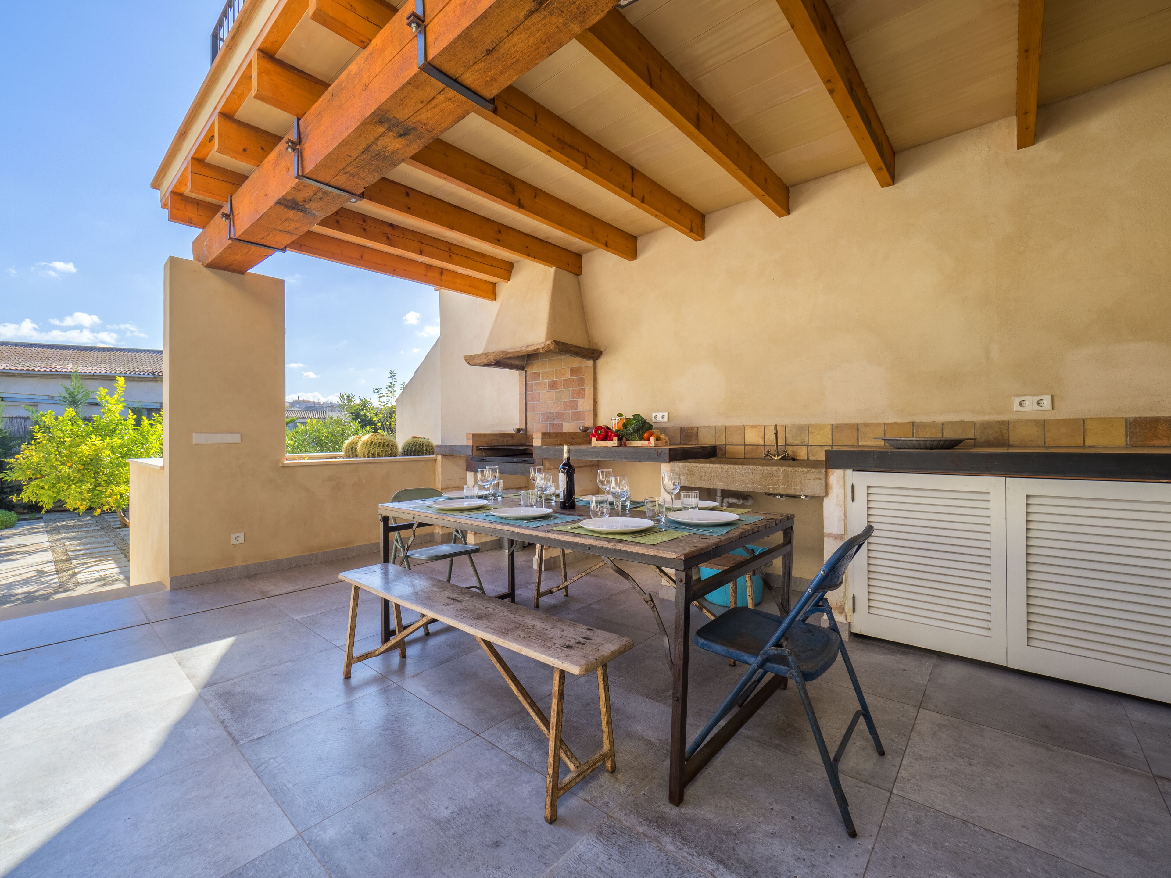 An outdoor patio with a wooden table set for dining, a bench, and a kitchen area with a brick oven.