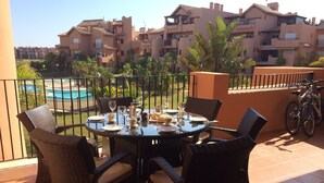 Un comedor con sillas de mimbre y una mesa redonda para cuatro, con vistas a una piscina y un edificio residencial.