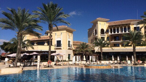 Un área de piscina con sillones de playa y sombrillas, palmeras y un edificio con varios balcones y ventanas.