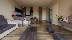 A modern living room with a sofa, ottomans, and a coffee table. There is a kitchen area with a TV and a dining table with chairs. - 3-star vacation rental. Large private balcony. Ocean View—Parking—Beach Access (Cassis)