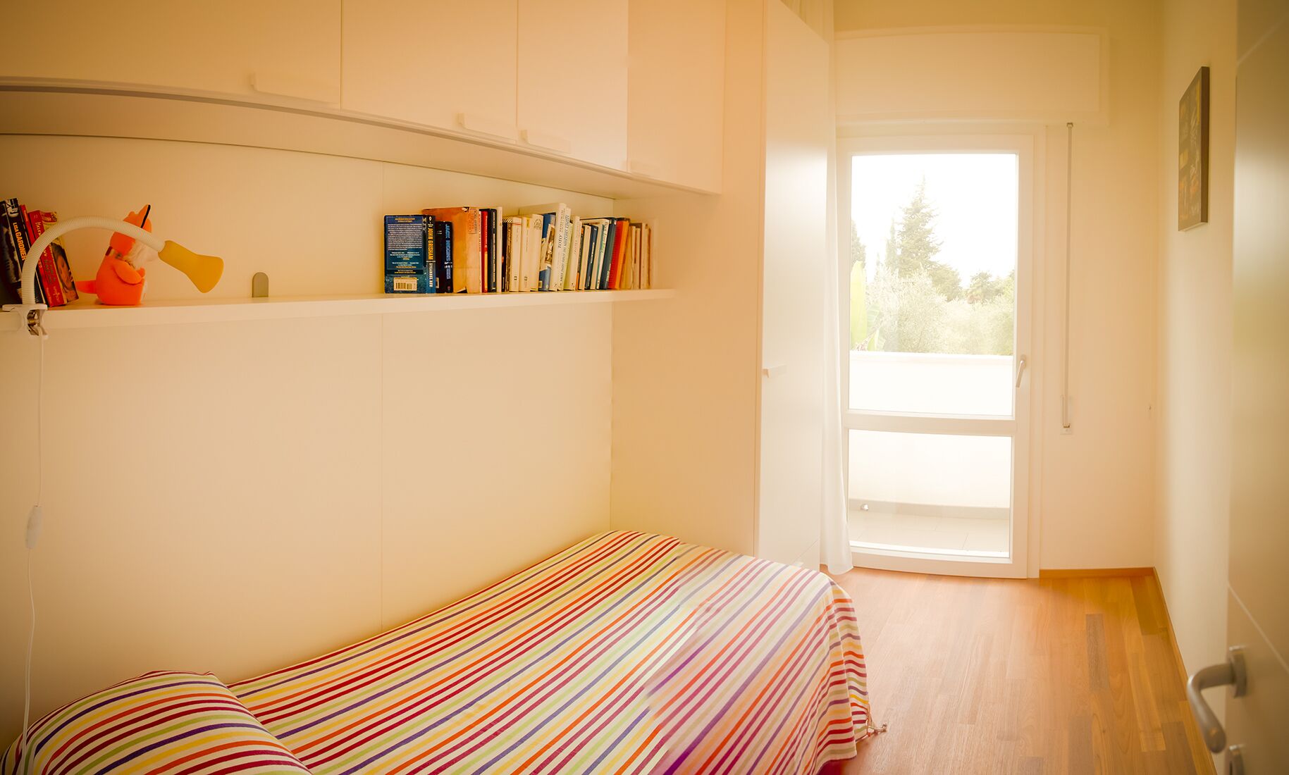 Une petite pièce lumineuse, avec un lit, une étagère remplie de livres et une porte donnant sur un balcon.