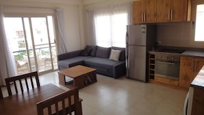 A compact living space with a sofa, a dining table, a kitchen area with a refrigerator and oven, and a sliding glass door leading to an outdoor area.