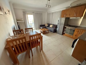 A modern living room with a dining area, a sofa, a kitchen with wooden cabinets, and a built-in oven.