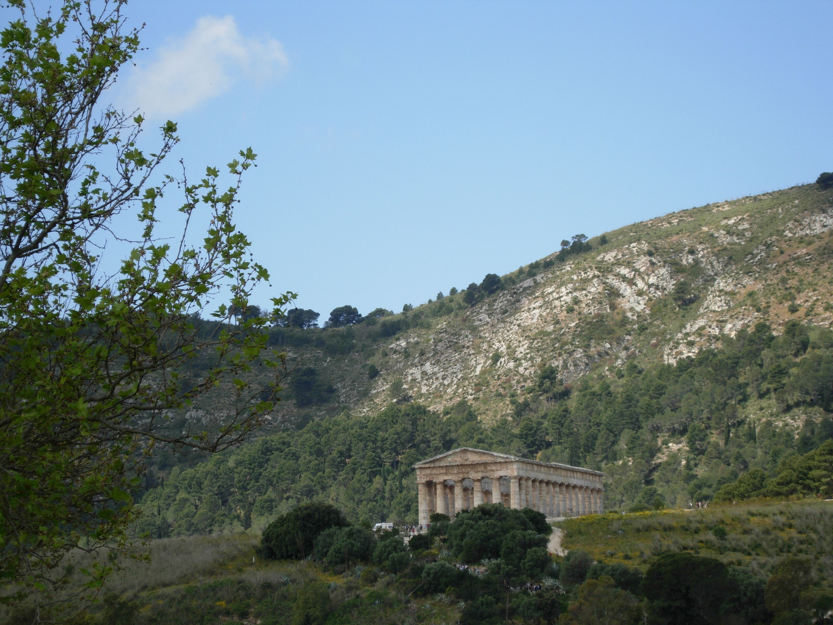 A classical building with columns, surrounded by a hilly landscape with trees and a clear sky.