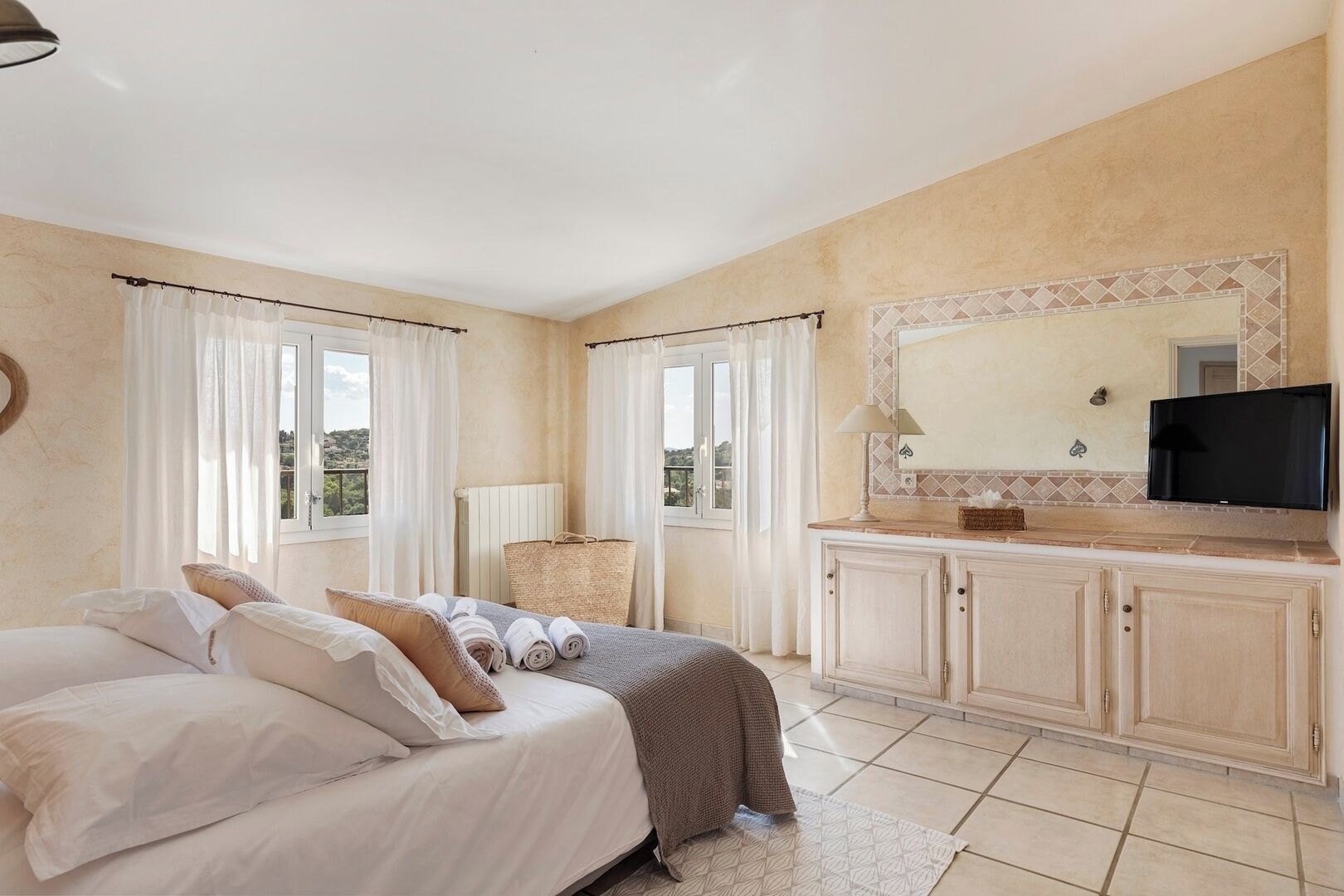 A bedroom with a bed, a vanity with a mirror, a television, and a view of the outdoors.