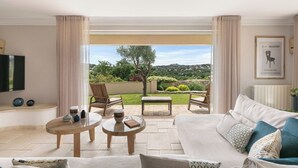 Ein Wohnzimmer mit einer Couch, zwei Sesseln, einem Couchtisch und einem Fernseher. Im Hintergrund ist ein Garten und eine Hügelkette zu sehen.