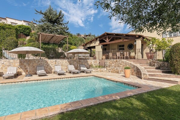 A swimming pool with lounge chairs and umbrellas, a stone wall, and a building with a clock.