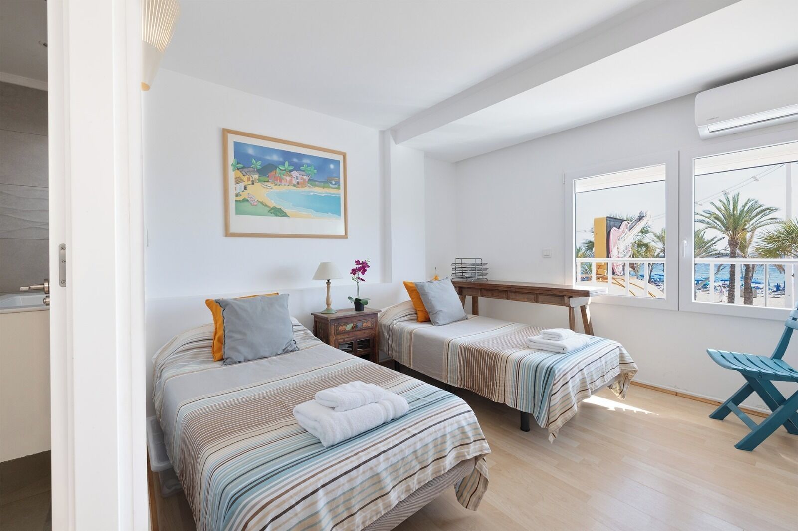 Une chambre avec deux lits, une petite table, une chaise, une fenêtre donnant sur la plage et un tableau au mur.