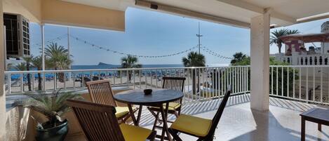 A balcony with a table and chairs, overlooking a beach with umbrellas and palm trees.