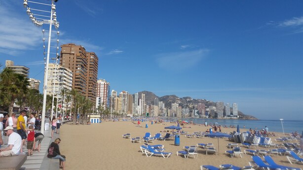 A beach with blue lounge chairs and umbrellas, a crowd of people, and tall buildings in the background. - Flat in Benidorm Renovated Frontline with Terrace (Benidorm)