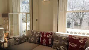 A cozy living room with a sofa, patterned cushions, a lamp, and a view of the city through large windows.