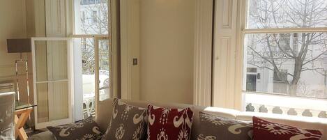 A cozy living room with a sofa, patterned cushions, a lamp, and a view of the city through large windows.
