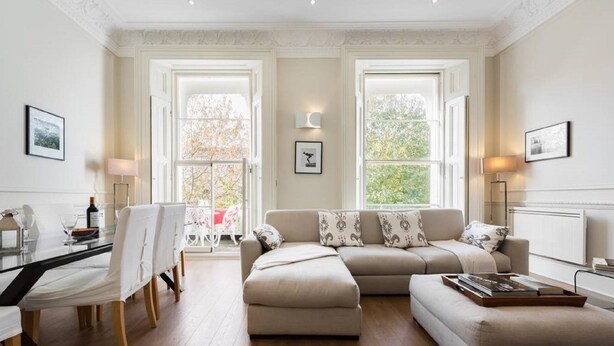 A spacious living room with a large beige sofa, a dining table with chairs, and two large windows with a view of trees.