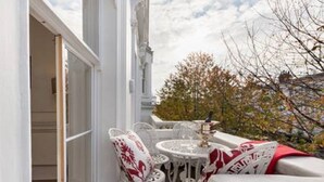 A balcony with white furniture, including chairs and a table, and a view of trees and buildings in the background.