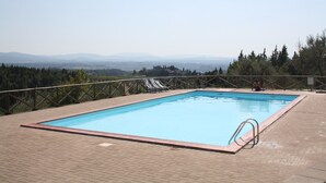 Une piscine rectangulaire avec un escalier en métal sur un côté, entourée d’une terrasse en briques. On y voit deux chaises longues et une personne assise au bord de la piscine. La piscine est située devant un décor composé d’arbres et de montagnes lointaines.
