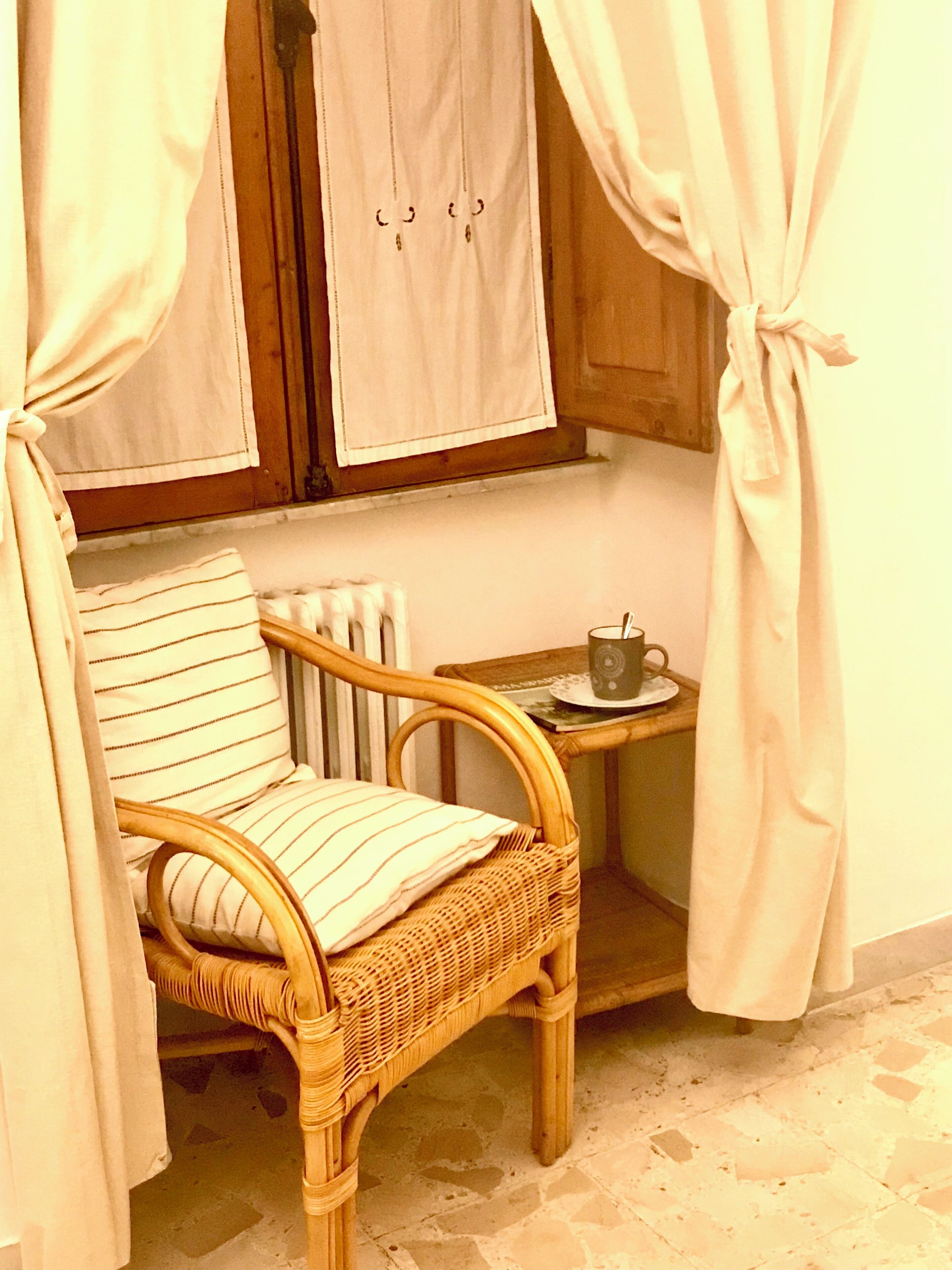 A wicker chair with a striped cushion, a small table with a cup, and a window with curtains.
