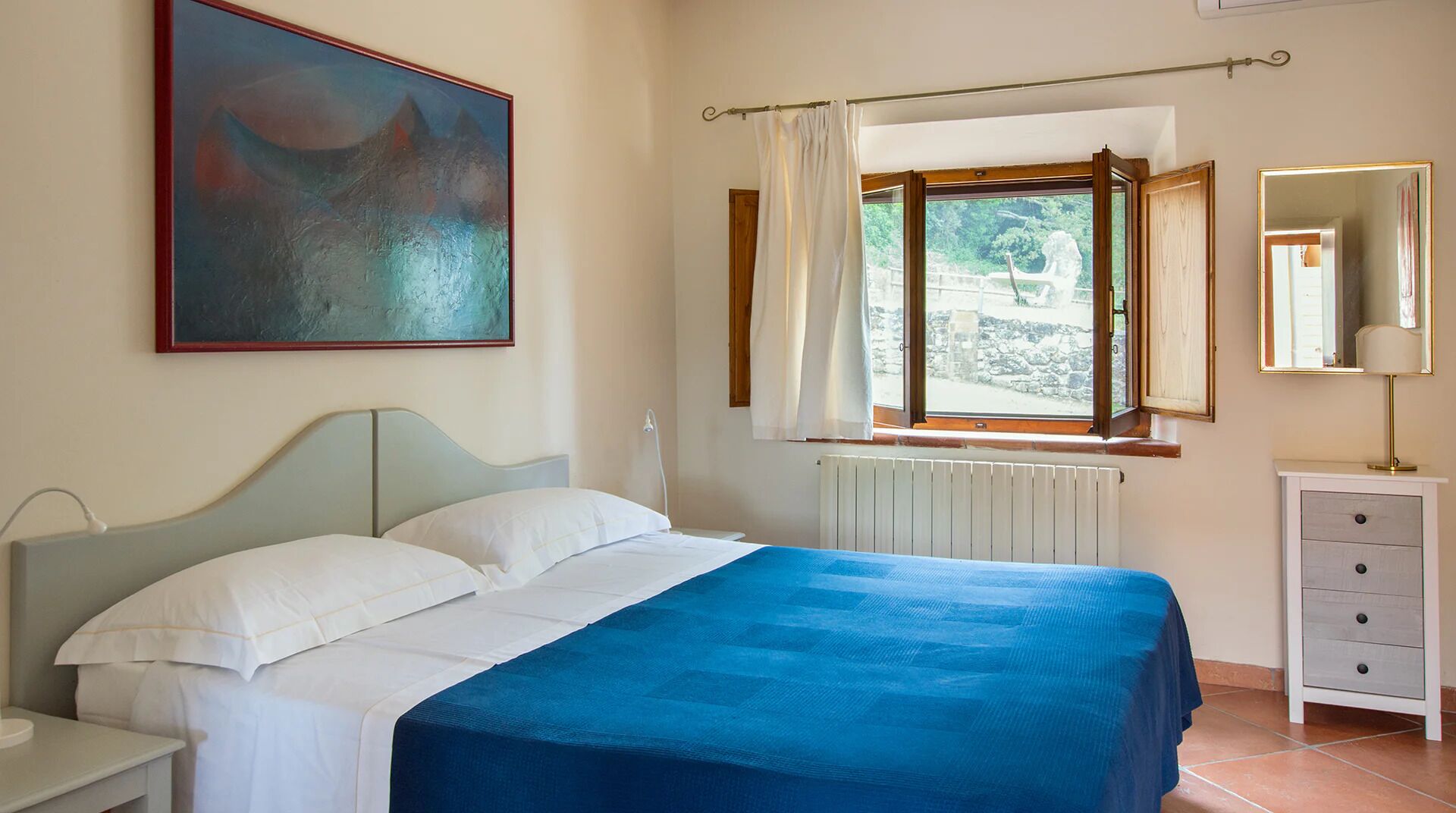 A bedroom with a bed, a nightstand, a dresser, a window with curtains, a radiator, and a painting on the wall.