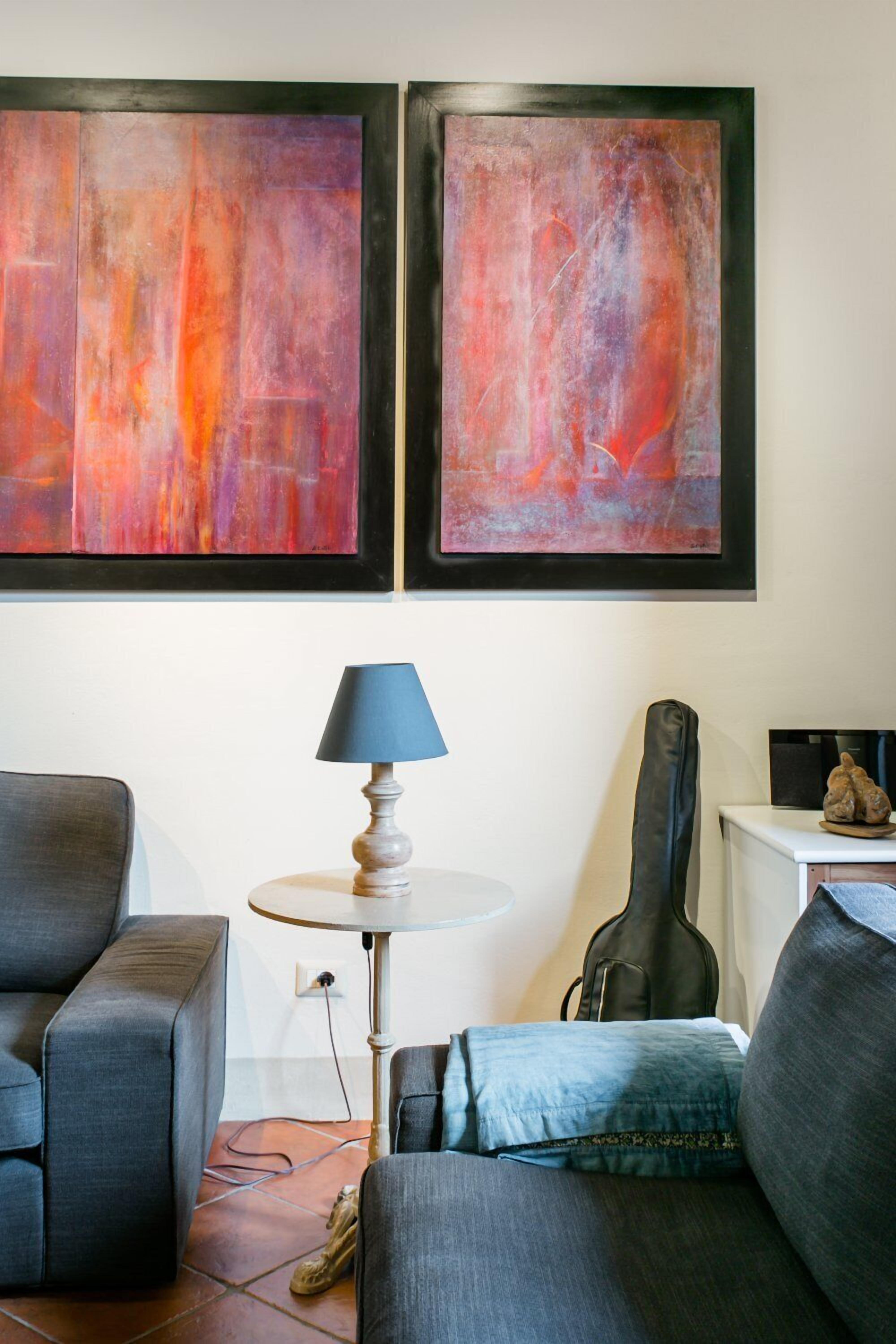 Una sala de estar con un sofá gris, una pequeña mesa redonda con una lámpara y dos cuadros abstractos en la pared.