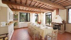 Un comedor con una mesa larga dispuesta para cuatro personas, vigas de madera en el techo, una chimenea y vistas al exterior con vegetación.