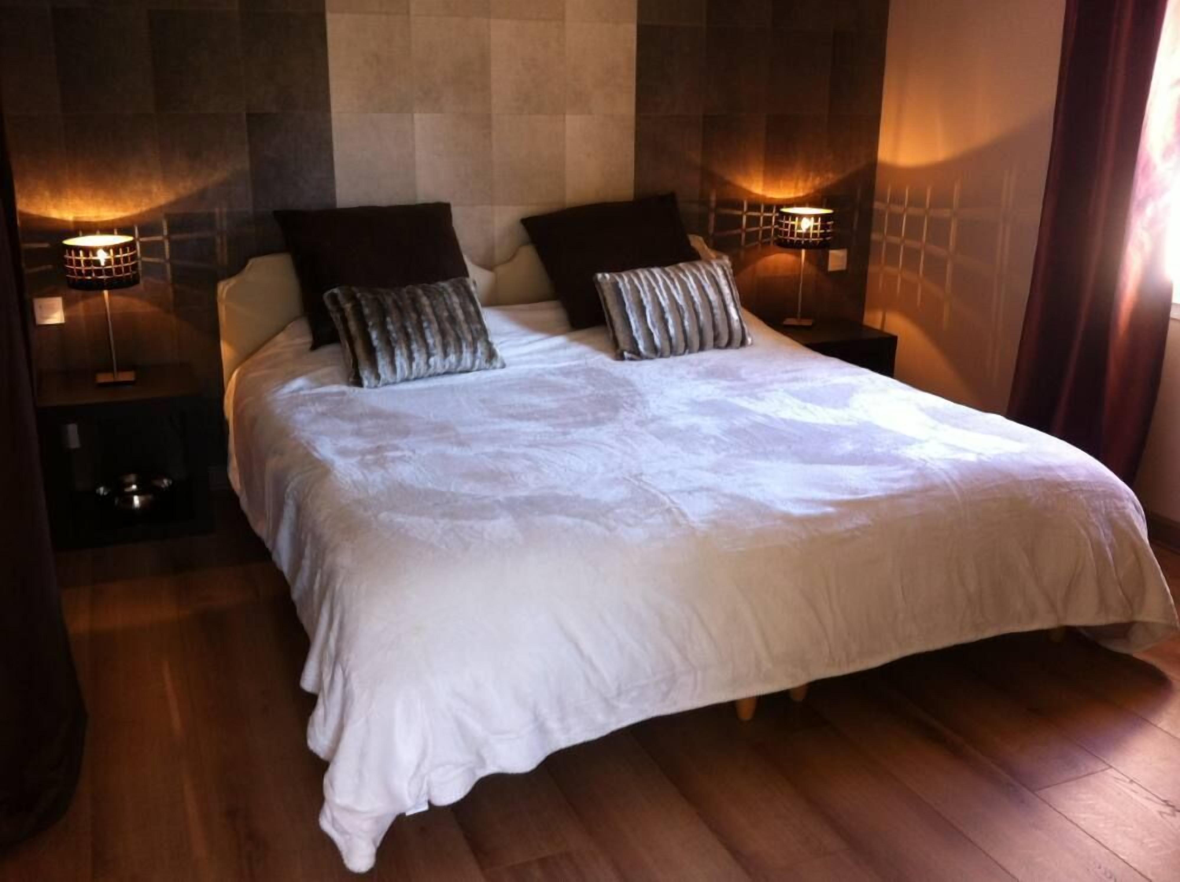 Una cama doble con edredón blanco, dos almohadas oscuras y dos lámparas de noche.