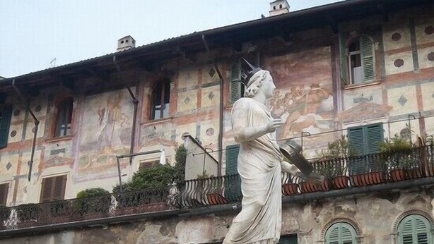 Une statue d’une femme, représentée dans une pose classique, se dresse sur un piédestal devant un bâtiment doté de nombreuses fenêtres et de balcons.