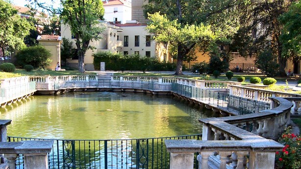 Um lago circular com uma ponte, rodeado por balaústres de pedra e vegetação.