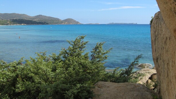 Vista costeira com mar azul límpido, pedras em primeiro plano e colinas ao longe.