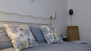 Uma cama com duas almofadas, uma mesa de cabeceira, uma luminária na parede e uma cesta de palha.