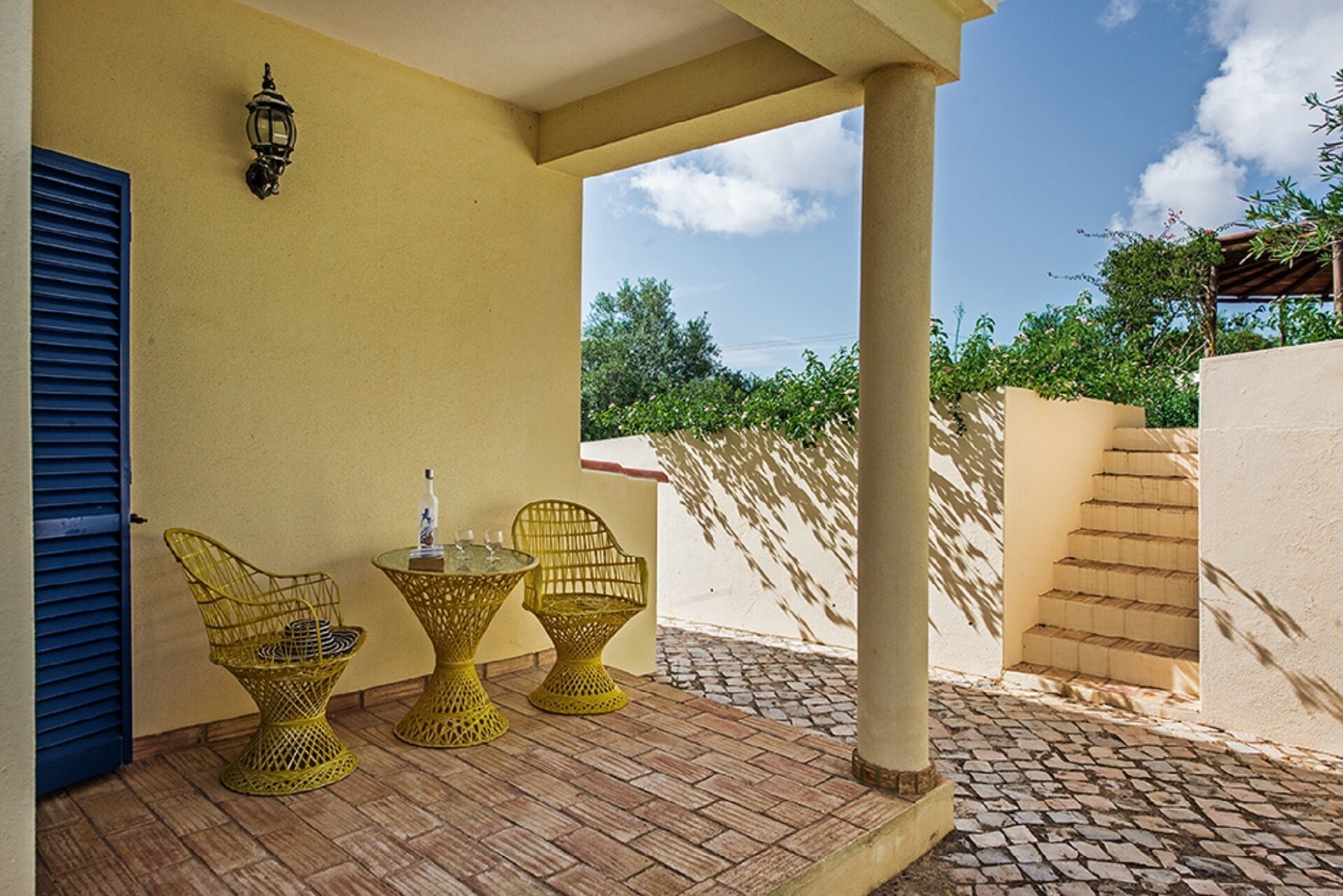 A covered patio with wicker chairs and a glass table, a bottle, and a wall-mounted lamp.