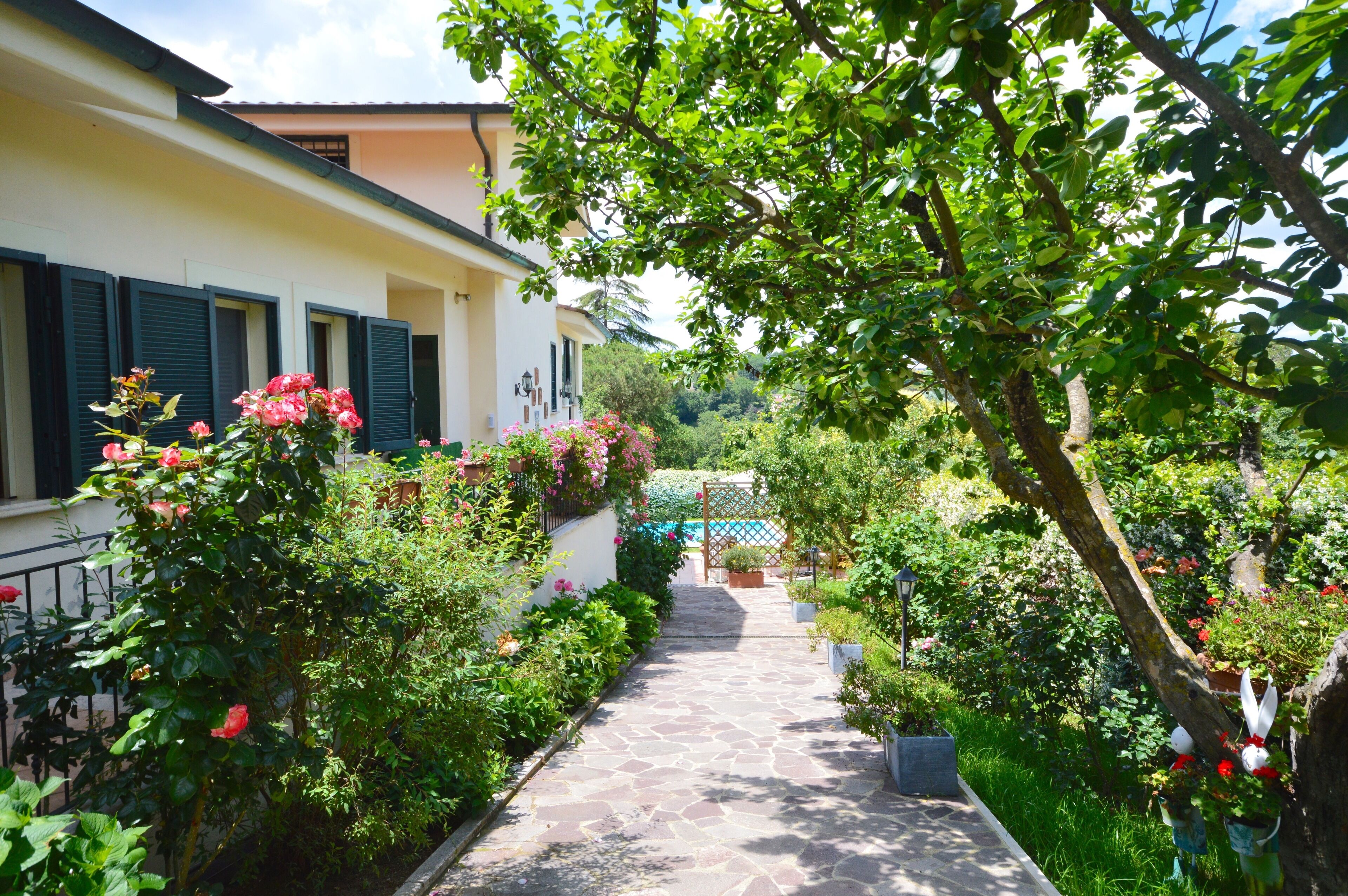 Un vialetto in pietra conduce a un edificio con persiane, immerso nel verde e fiorito.
