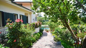 Un vialetto in pietra conduce a un edificio con persiane, immerso nel verde e fiorito.