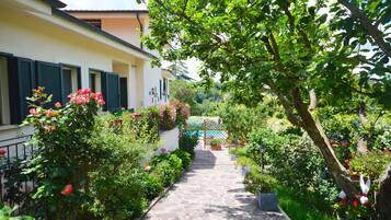 Un vialetto in pietra conduce a un edificio con persiane, immerso nel verde e fiorito.