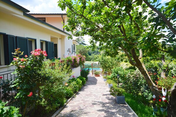 A stone pathway leading to a building with shutters, surrounded by lush greenery and flowering plants. - House / Villa - Rignano Flaminio (Rignano Flaminio)