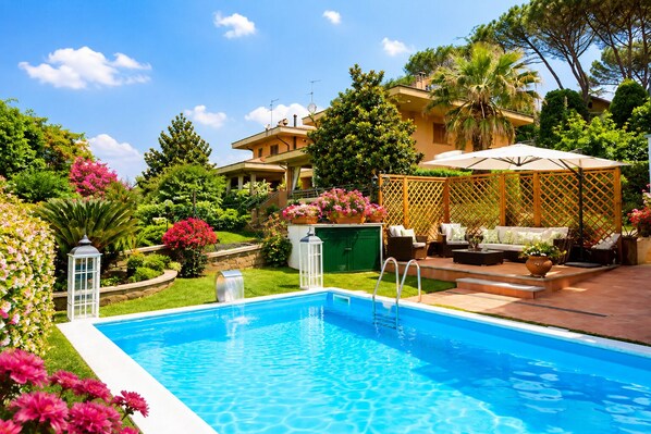 A swimming pool with a wooden deck and outdoor furniture, surrounded by lush greenery and a house. - House / Villa - Rignano Flaminio (Rignano Flaminio)