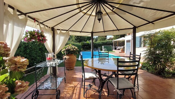 Un patio coperto con tavolo e sedie, un cesto di fiori appeso e vista su una piscina sullo sfondo.