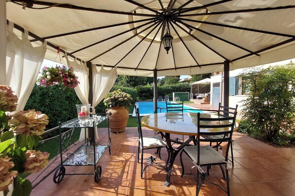 A covered patio area with a table and chairs, a hanging flower basket, and a view of a swimming pool in the background. - House / Villa - Rignano Flaminio (Rignano Flaminio)