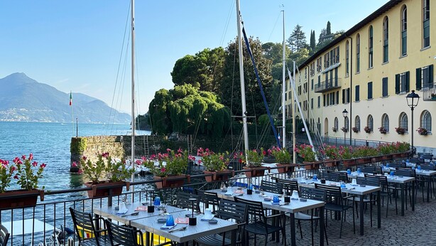 Ein Restaurant am Wasser mit Tischen, Stühlen, Pflanzen in Töpfen und Blick auf das Meer und die Berge.