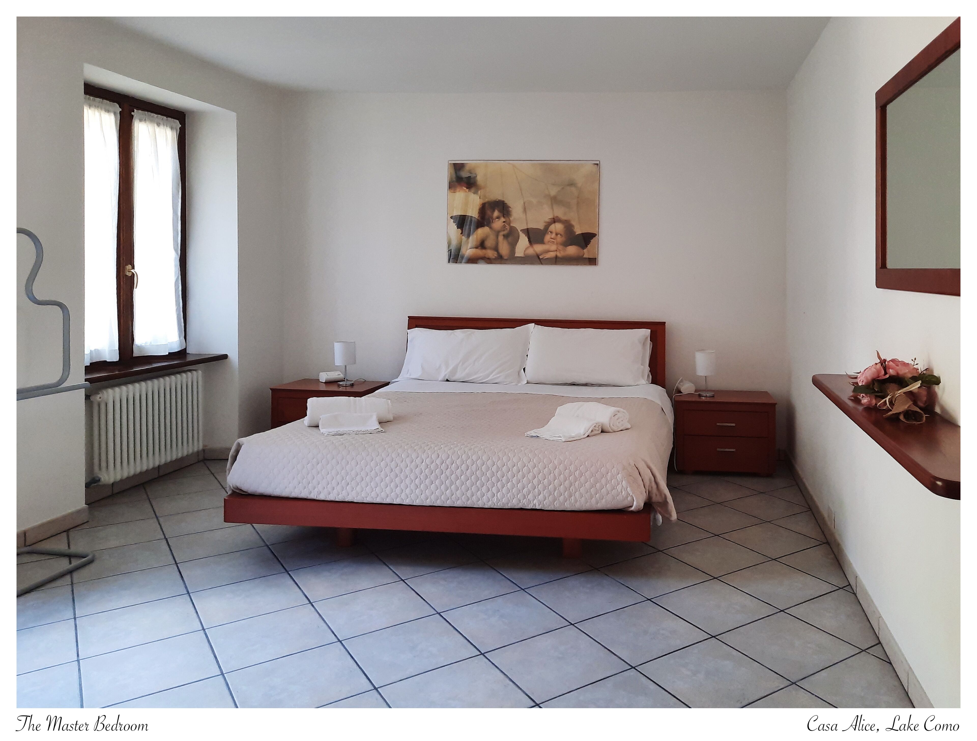 Une chambre principale dotée d’un grand lit, d’un radiateur, d’une fenêtre avec des rideaux, d’un tableau accroché au mur et d’une table de chevet avec une lampe.