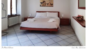 Ein Schlafzimmer mit großem Bett, Heizkörper, einem Fenster mit Vorhängen, einem Bild an der Wand und einem Nachttisch mit Lampe.