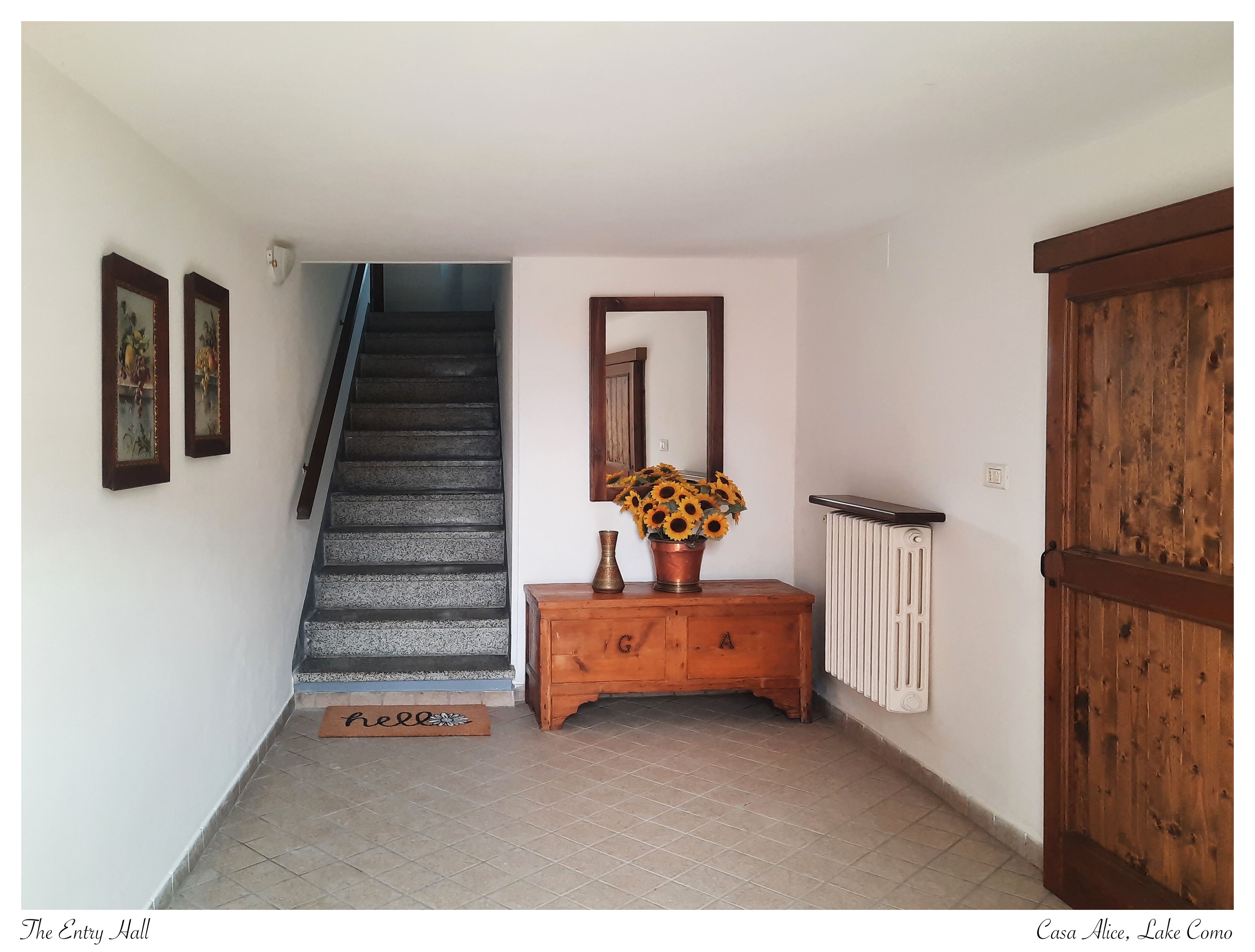 Ein Korridor mit einer Holztreppe, einem Konsolentisch aus Holz mit einer Vase Sonnenblumen, einem Heizkörper und einer großen Holztür.