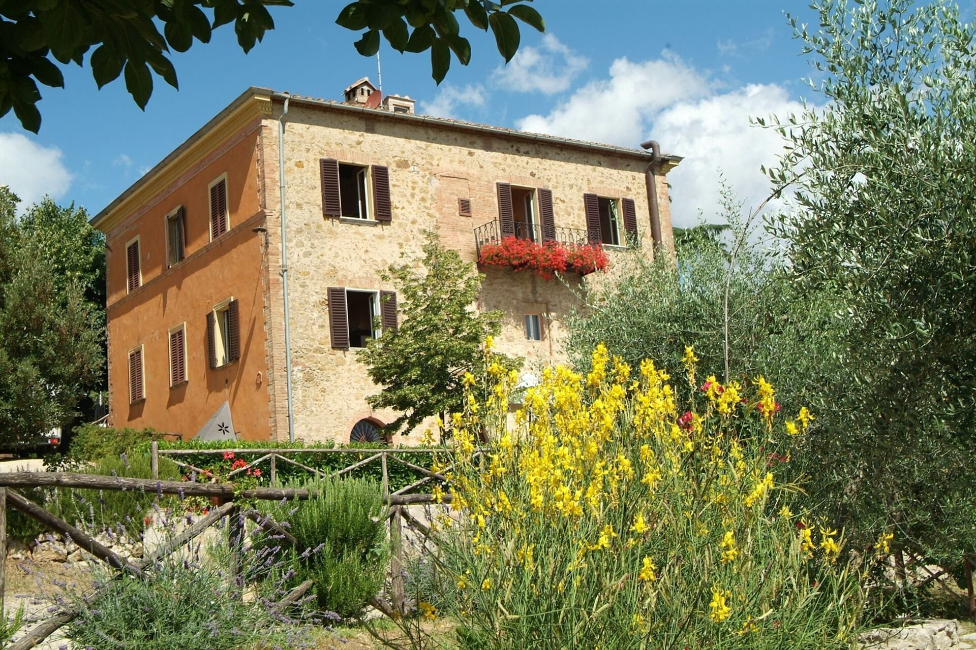 A two-story building with a stone facade, multiple windows with shutters, and a balcony with red flowers.