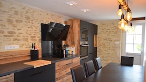 A modern kitchen with a wooden island, built-in oven, and a dining area with a dark wooden table and chairs. - Charming house, enclosed garden, 3 min from Arromanches and the beaches (Ryes)
