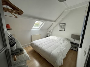 Une chambre à coucher avec un grand lit, une lucarne, un radiateur, une table de chevet avec une lampe et un tableau encadré au mur.