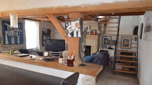 A rustic interior with a wooden counter, stone pillar, and a staircase leading to a loft area. - Charming house, enclosed garden, 3 min from Arromanches and the beaches (Ryes)
